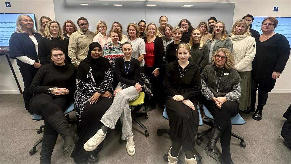 Ett trettiotal personer står i klassrum med en whiteboardtavla i bakgrunden och tv-skärmar på vardera sida. 