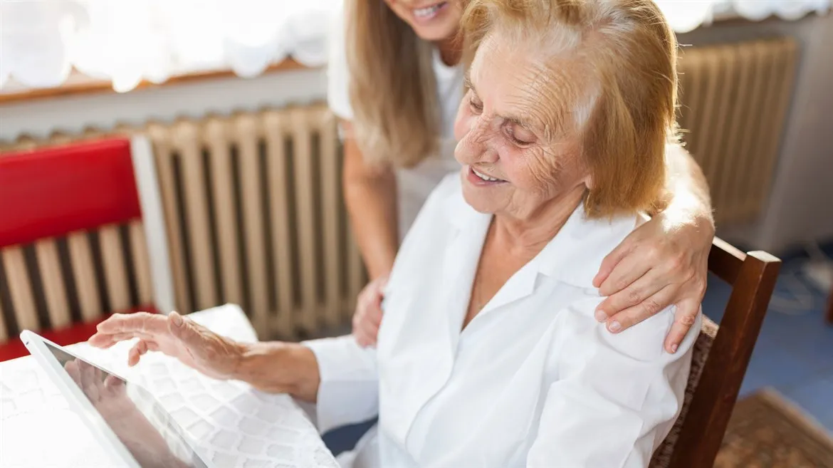 Äldre kvinna tittar på en tablet, yngre kvinna står bakom och håller henne om axlarna.