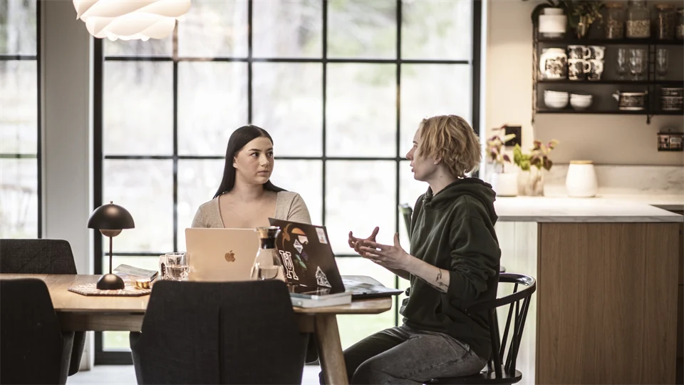 Två kvinnor sitter vid ett matbord och jobbar framför varsin dator. 