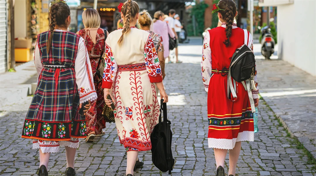 Tre kvinnor i traditionella bulgariska folkdräkter går på en gata i en stad. Dräkterna är färglada med starka färger och mönster. 