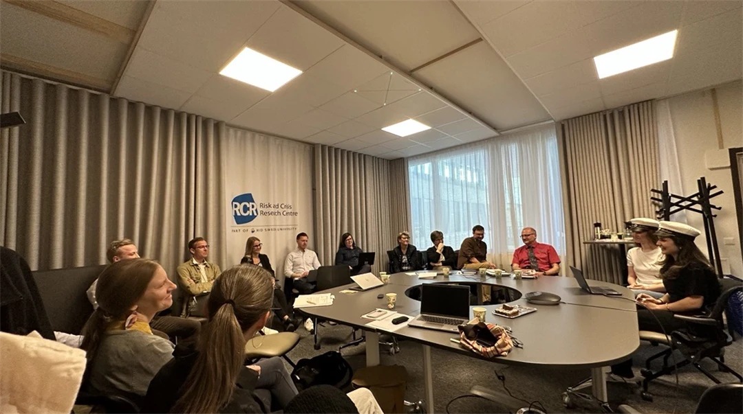 Fyra personer sitter runt ett ovalt konferensbord med laptops, papper och kaffekoppar. En skylt med texten "Risk and Crisis Research Centre" syns på väggen bakom dem.