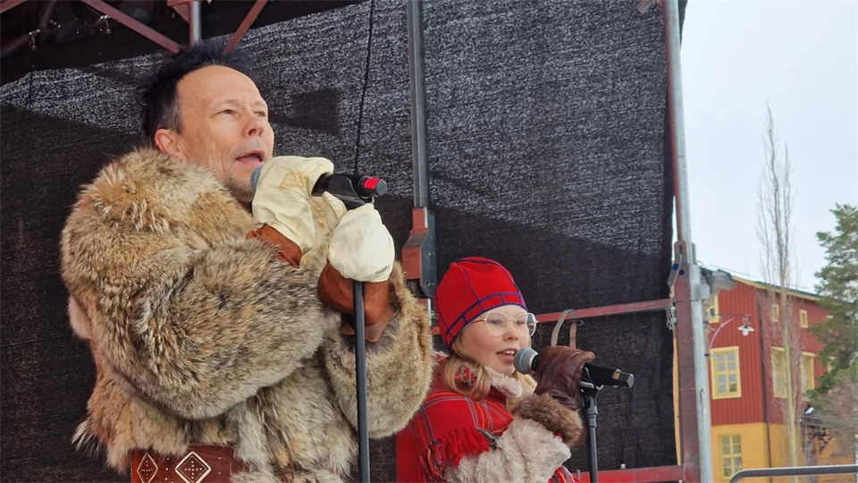 En man och en flicka i pälsar sjunger på en scen.