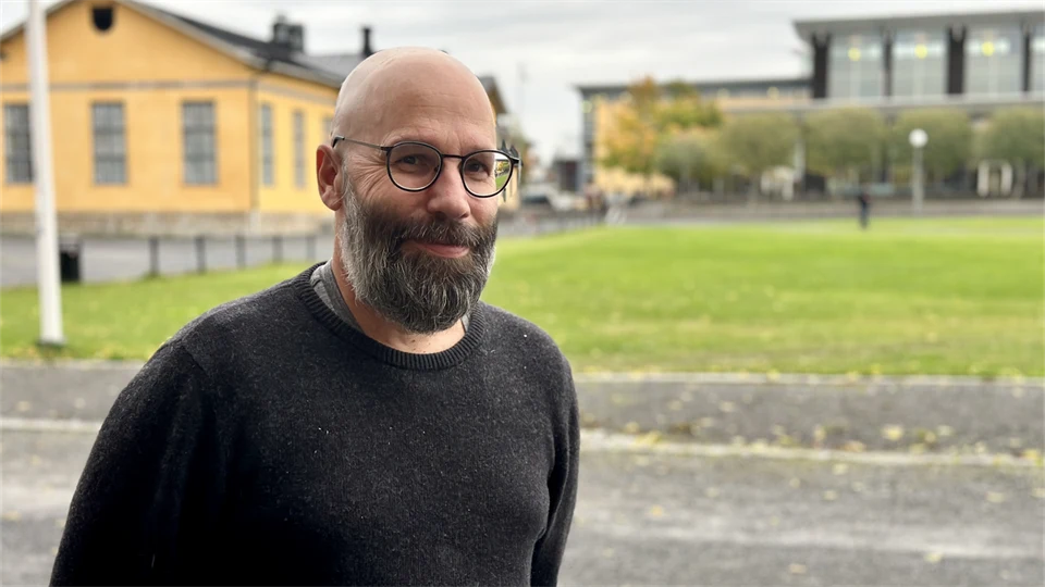 En man med glasögon och en mörkgrå tröja som står utanför framför gula byggnader och ler in i kameran