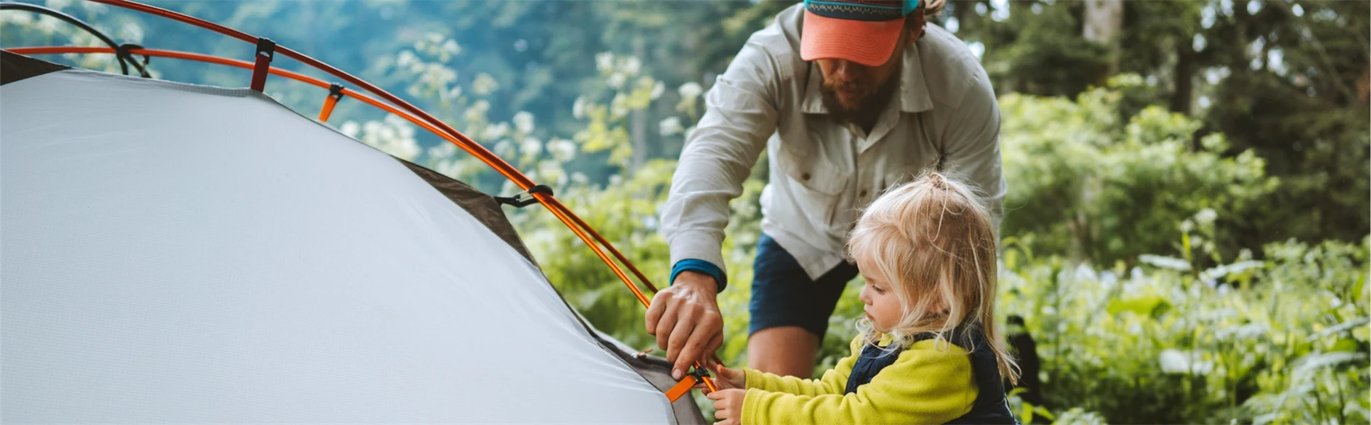 Ung man och hans dotter tre-årsåldern sätter upp ett tält tillsammans i skogen. De campar och är klädda i friluftskläder.