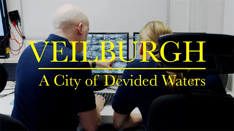 Två personer sitter vid ett skrivbord och tittar på en datorskärm med flera övervakningsbilder. På bilden finns en stor gul text som lyder: ”VEILBURGH – A City of Devided Waters”.