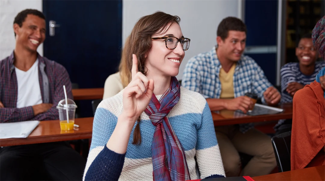 Kvinnlig student sträcker upp handen i ett klassrum där hon sitter vid en bänk tillsammans med andra studenter