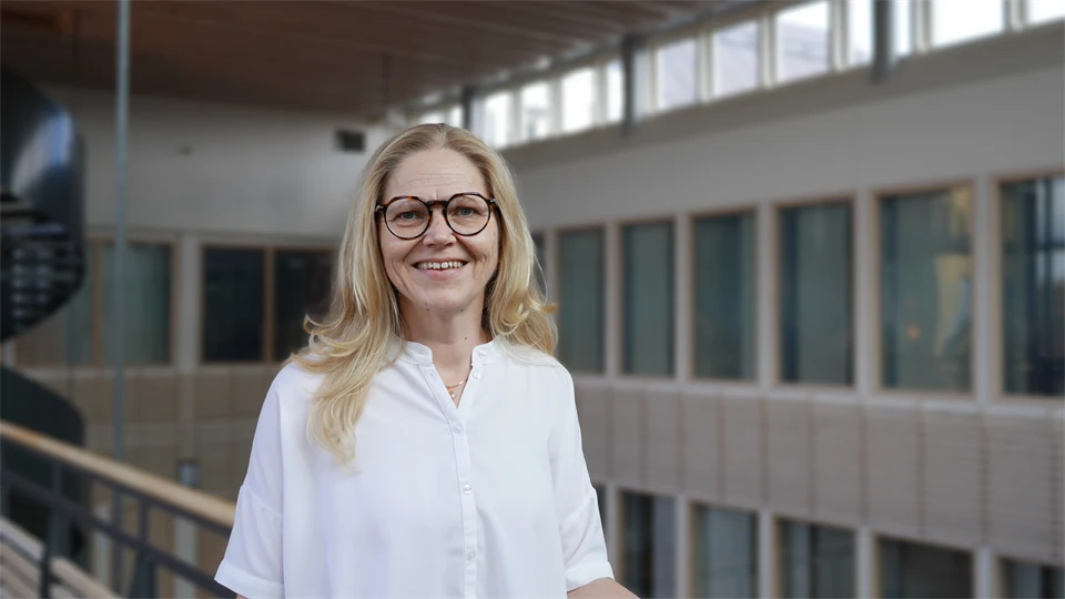 En ljushårig kvinna i vit blus och glasögon ler mot kameran. Bakgrunden är suddig.