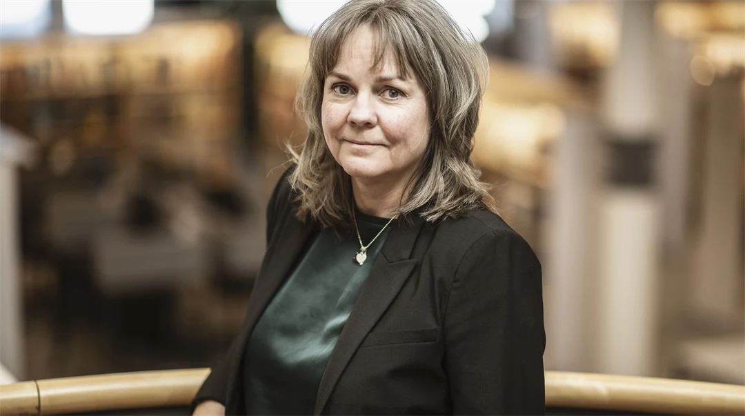 A woman with light brown hair, wearing a green blouse and black jacket, stands in a library setting.