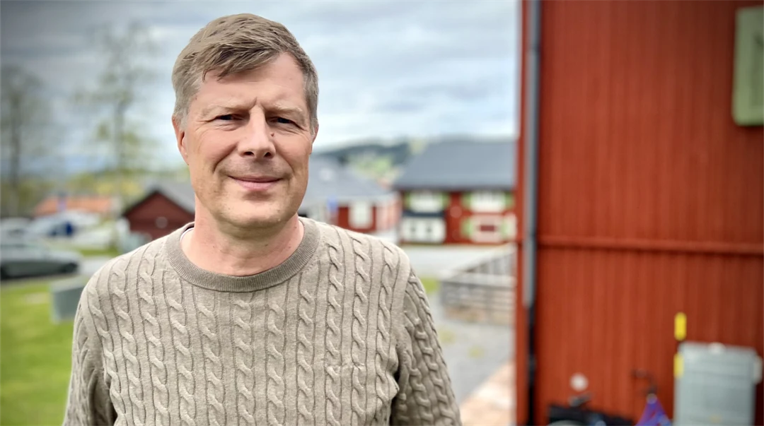 Man i beige tröja står utomhus med en röd husvägg i bakgrunden.