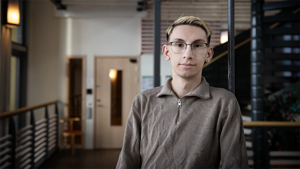 En man med glasögon och beige tröja står i en korridor.