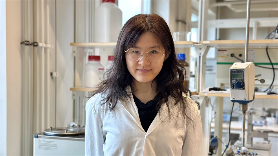 Young woman with long dark hair wearing a lab coat, standing in a laboratory.