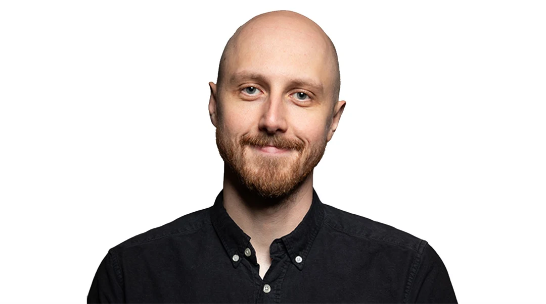 Man med skägg och mustasch ler mot kameran, klädd i svart skjorta, vitt bakgrund.