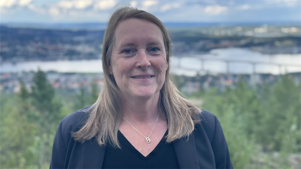 En kvinna med blont axellångt hår, svart tröja och mörkblå kavaj med en naturskön vy med skog, hav och en bro i bakgrunden.