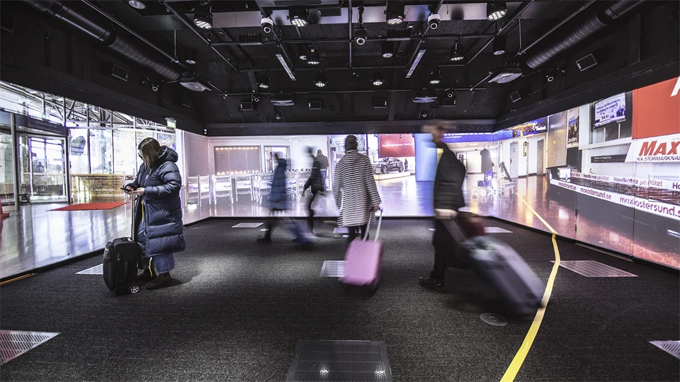 Several people are standing in a room with images from an airport projected on the walls. They have coats and suitcases. There is a yellow strip of tape on the floor. There are lots of technical equipment in the ceiling.