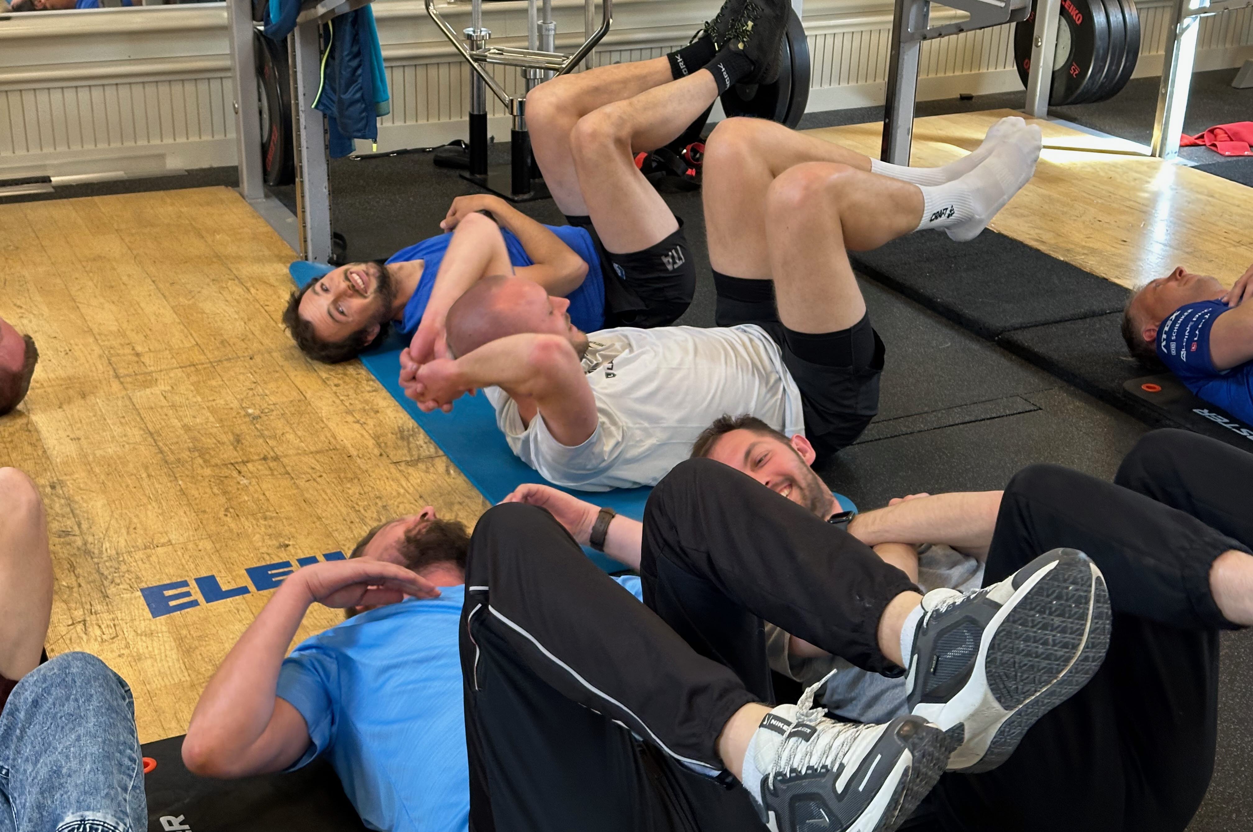 A group of men do a physical exercise lying on the floor.