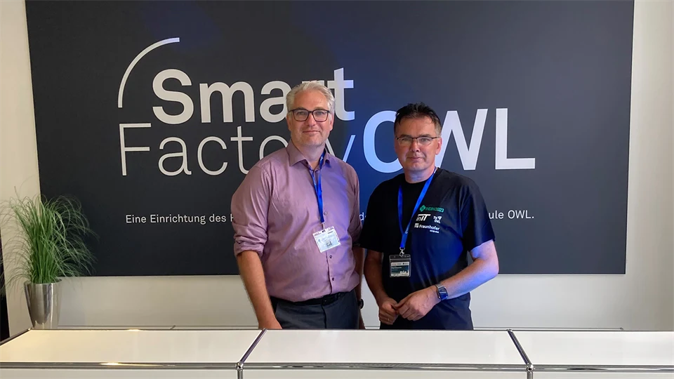 Two men stand and smile at the camera with a sign behind them that says Smart Factory OWL