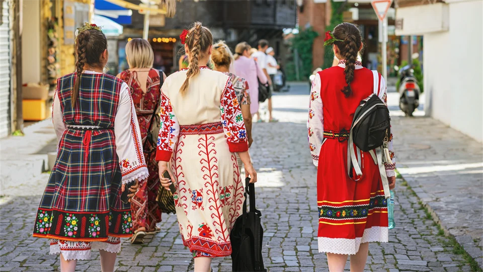 Three women in traditional Bulgarian folk costumes walk down a city street. The costumes are colorful with strong colors and patterns.