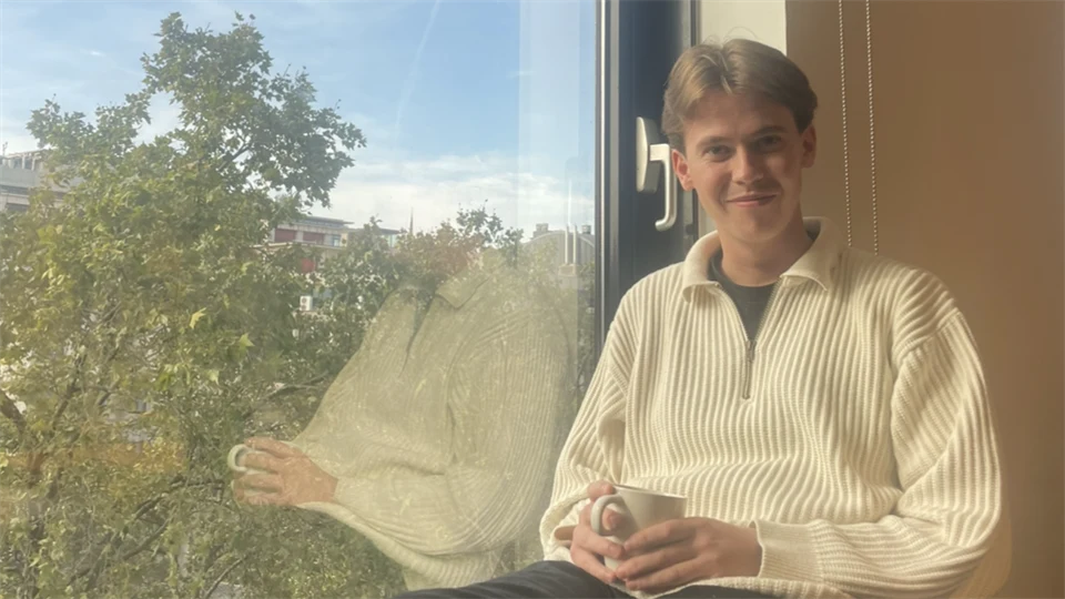 Ung man som sitter i ett fönster med en vit stickad tröja på sig och med en kaffekopp i handen. 