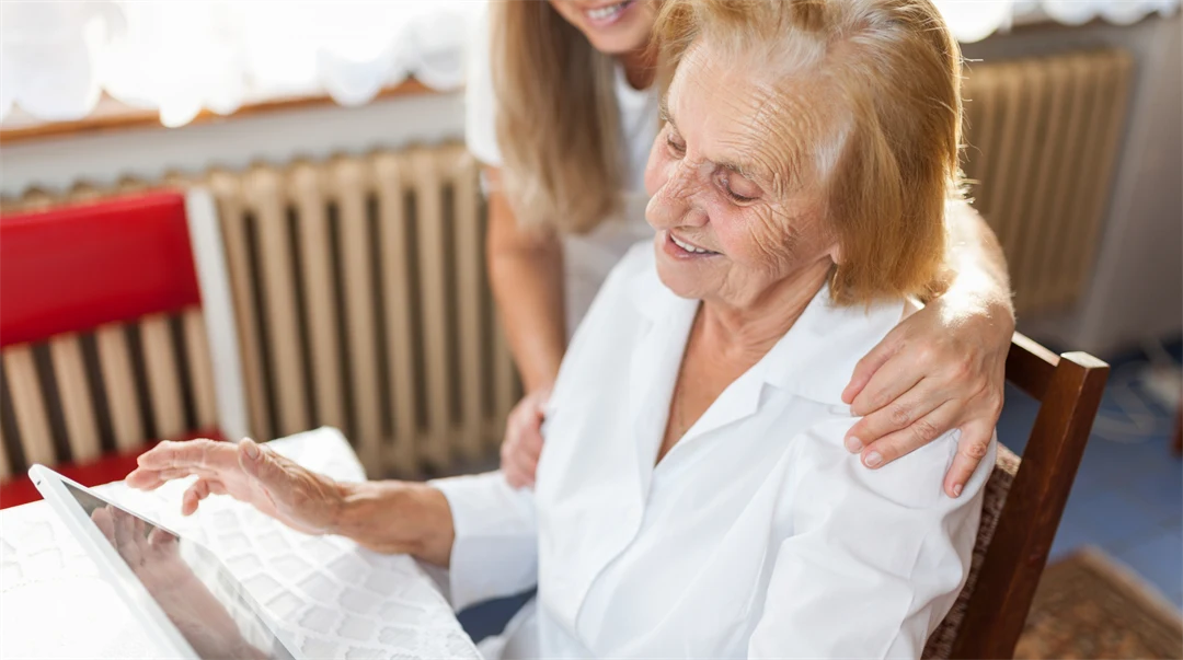 Äldre kvinna tittar på en tablet, yngre kvinna står bakom och håller henne om axlarna.