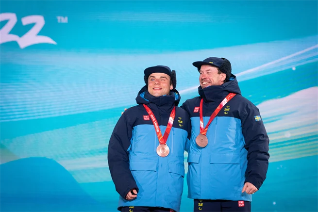 Två män klädda i klarblå jackor med marinblå ok och ärmar, samt marinblå kepsar står framför en blå fond med siffrorna 22 i övre vänstra hörnet. Båda män har en medalj med rött band runt halsen. De ser glada ut.