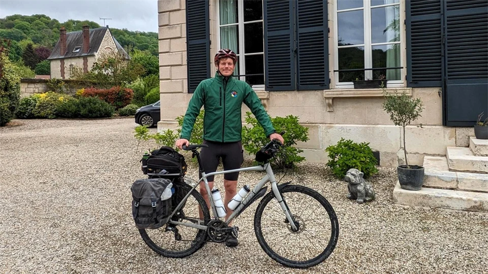 En man i grön jacka och röd cykelhjälm står framför ett hus med sin cykel framför sig