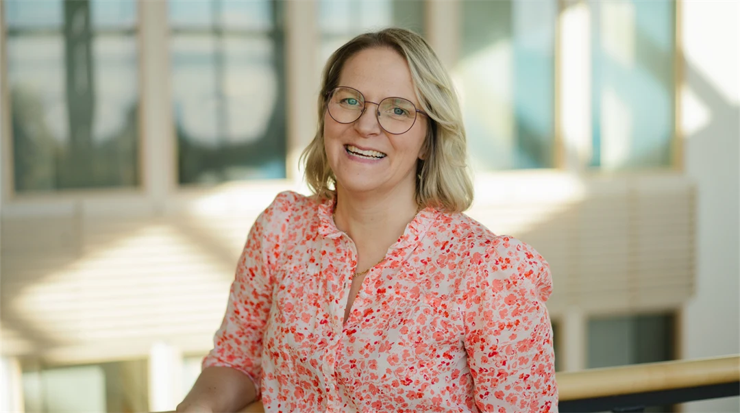 : Glad kvinna med blont hår, blommig blus och glasögon ler mot kameran. 