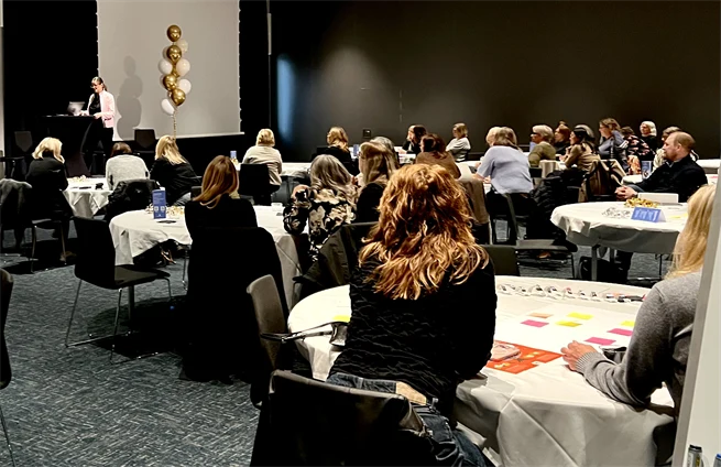 Ett rum med runda bord och svarta stolar med ett femtiotal deltagare som sitter ner vid borden. Längst fram i salen står en kvinna på en scen med guldiga och vita ballonger.