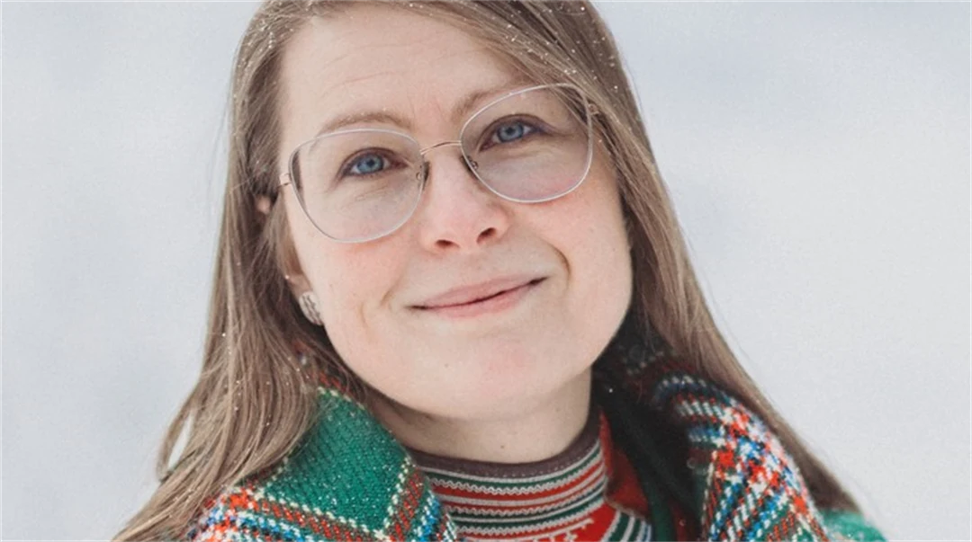 Woman with glasses and dark blonde hair with a chequered shawl over her shoulders looks at the camera.