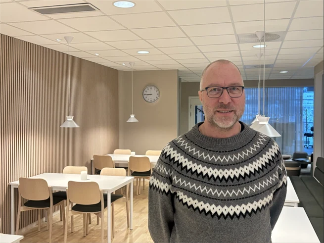 Man i grå stickad tröja med glasögon ler mot kameran, en sal med ljust interiör, stolar och vita bord i bakgrunden.