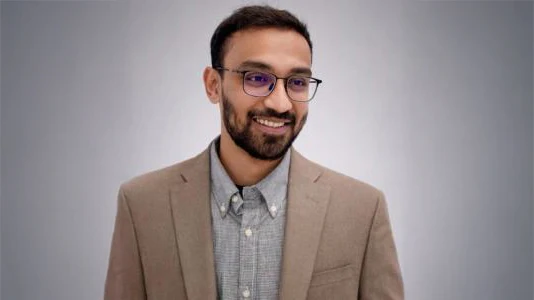 A smiling man with a beige jacket and glasses