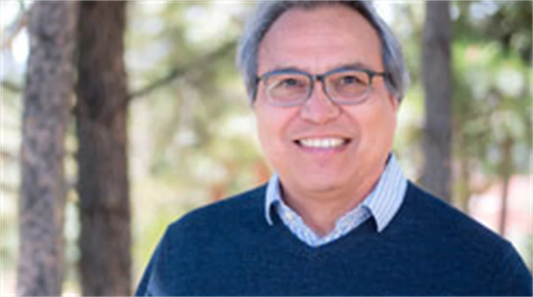 A man with glasses wearing a blue and white checkered shirt with a blue sweater over it stands outside with trees in the background