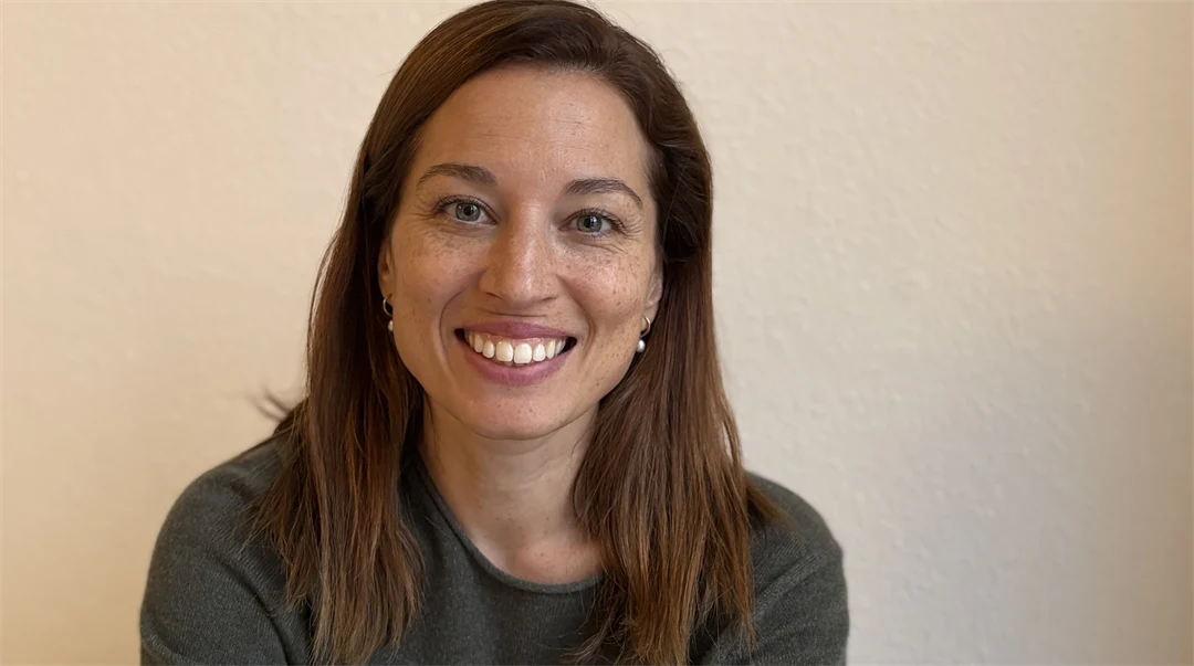 Kvinna med långt brunt hår med fräknar, ler mot kameran, klädd i grå tröja.