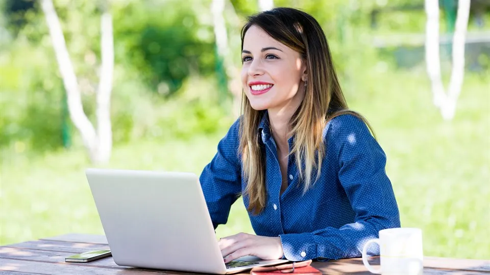Smiling young woman sitting outdoors in front of a laptop, notebook and a cup of coffee.