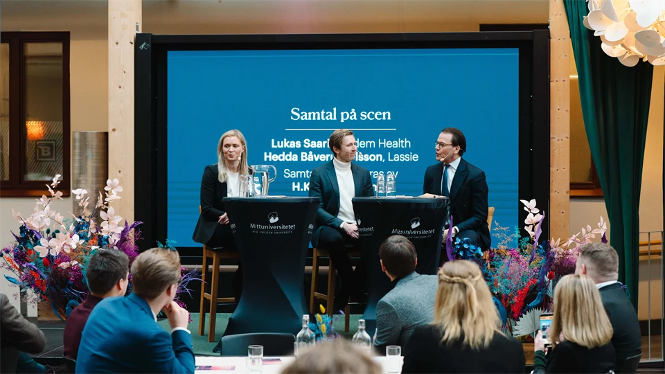 Two men and a woman are sitting on a stage with a large screen behind them. In front of them is an audience.