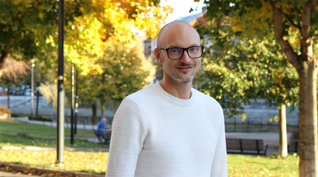 Man med glasögon och vit tröja poserar, träd med höstlöv i bakgrunden.