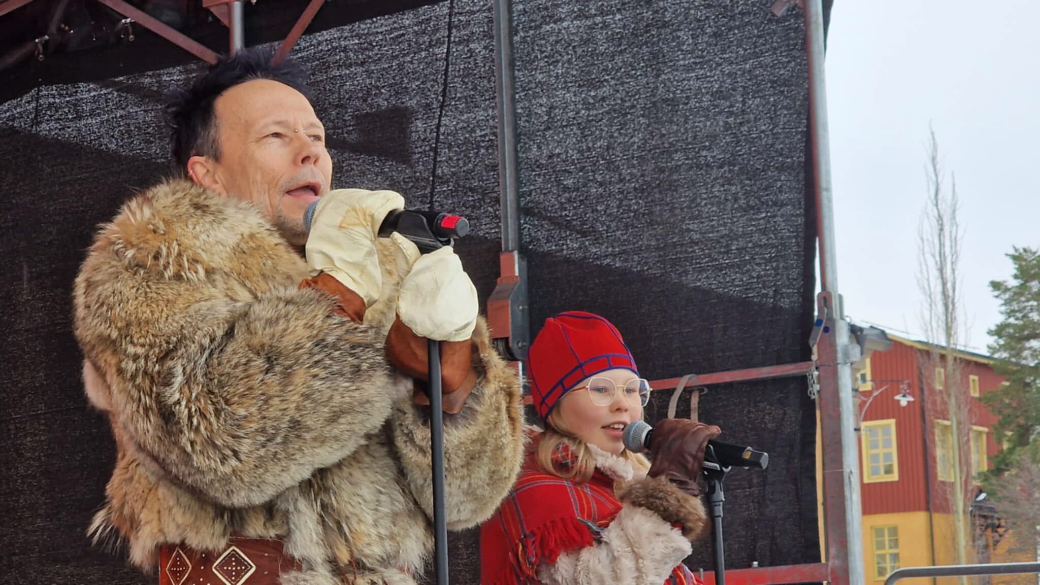 En man och en flicka i pälsar sjunger på en scen.