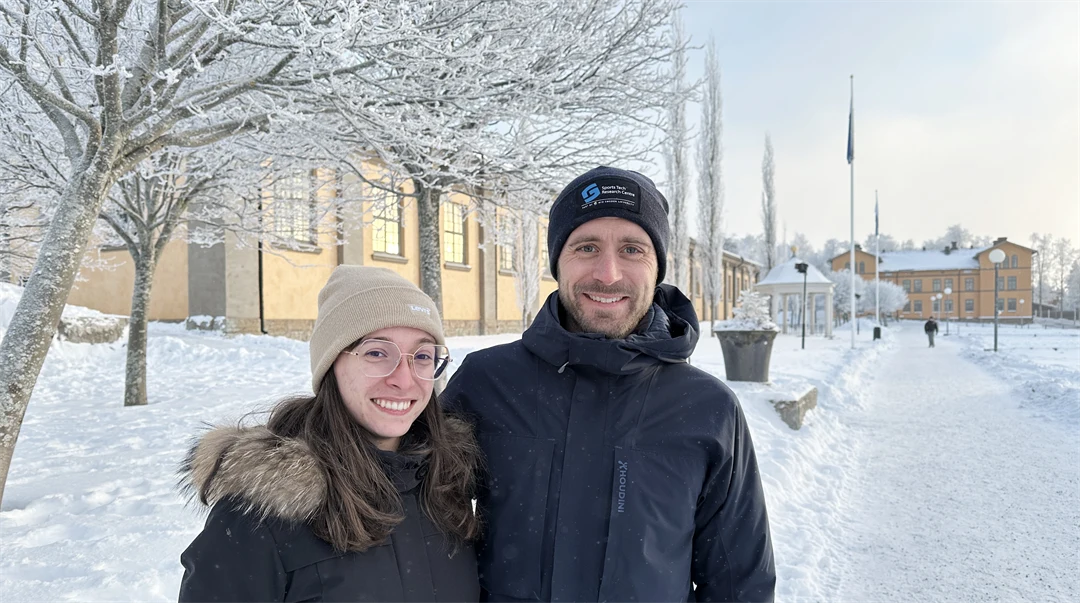 Två personer står framför frostiga träd och byggnader klädda i vinterkläder.