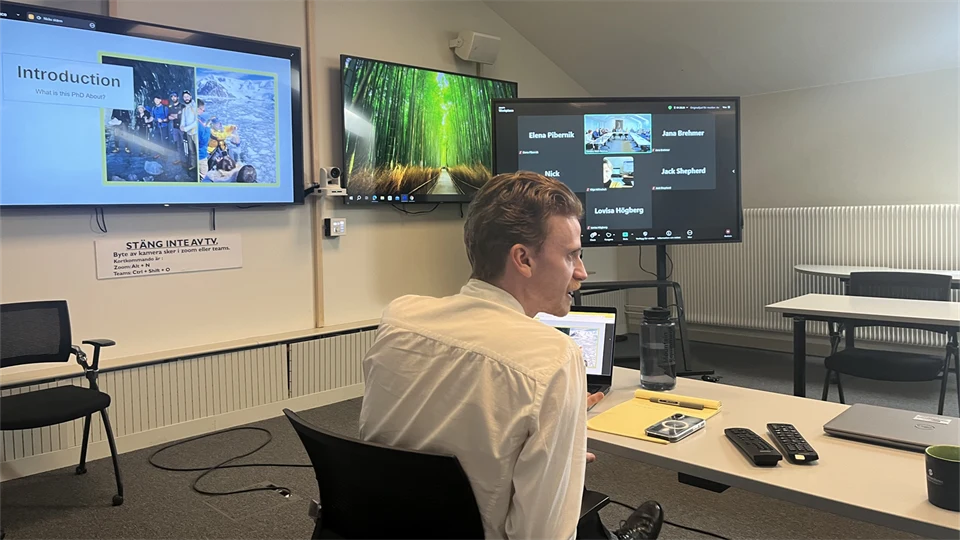 En man i vit skjorta sitter vid ett bord med en dator framför sig och i bakgrunden syns tre skärmar.