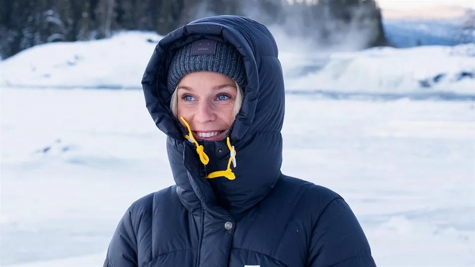 En kvinna i mörkblå täckjacka med huvan uppdragen och mössa står utomhus. Bakom henne finns ett snötäckt fält och längre bort granskog. Hon tittar åt sidan och ler. 