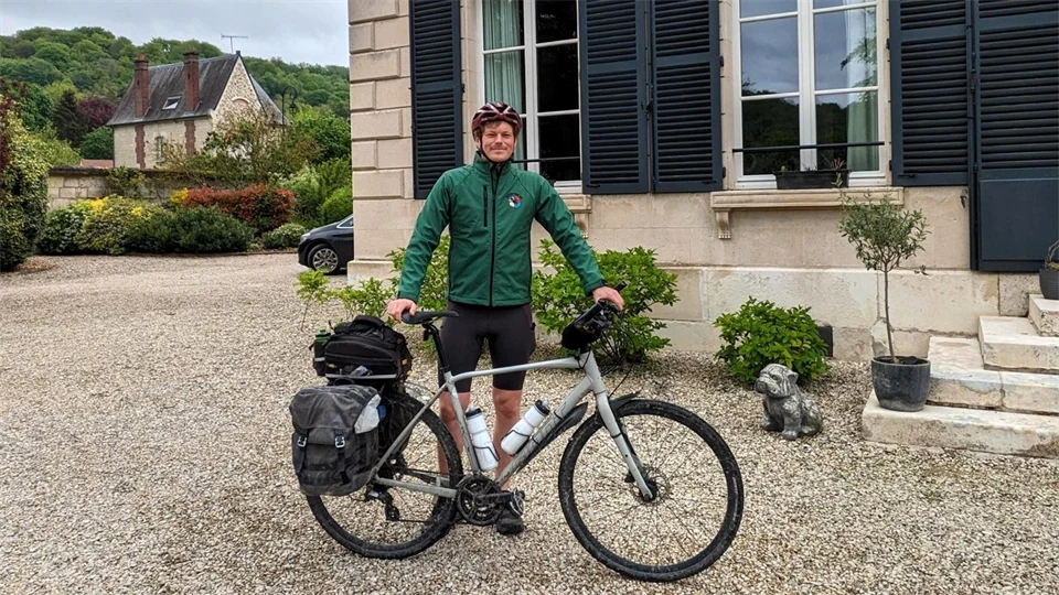 En man i grön jacka och röd cykelhjälm står framför ett hus med sin cykel framför sig