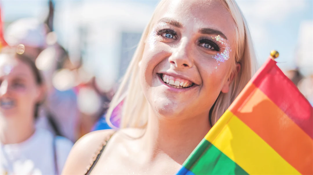 Leende kvinna med blont hår, glitter runt ögonen och en prideflagga i handen 