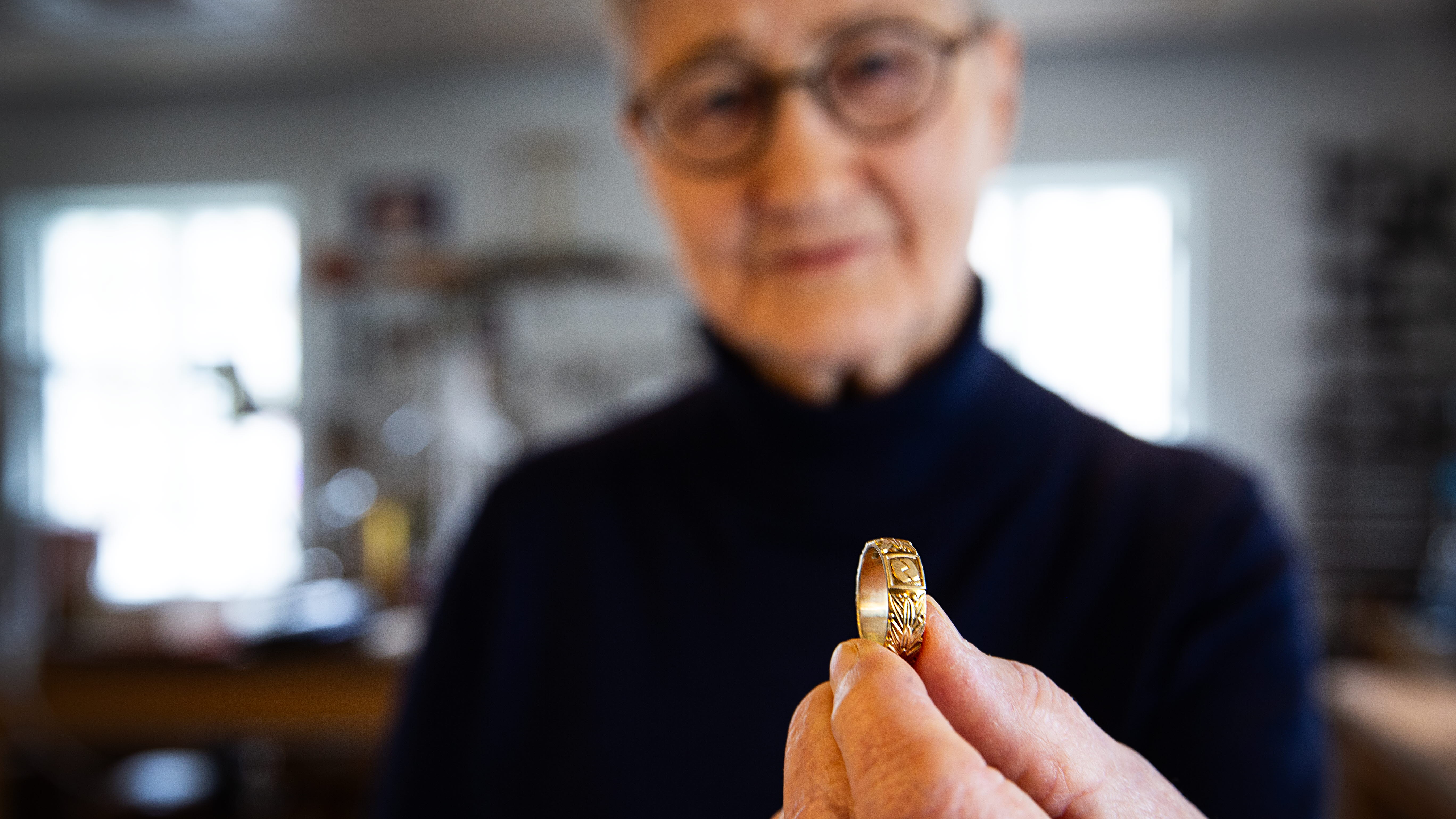 A woman with glasses and short hair is holding a gold ring in front of the camera. The focus is on the ring.
