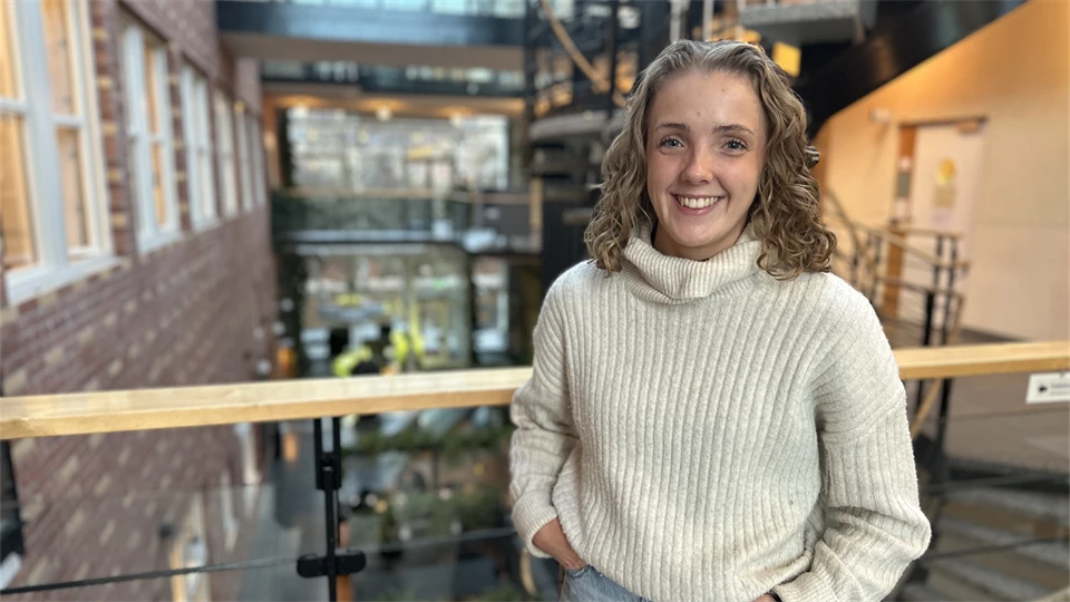 Ung kvinna med blont, lockigt, axellångt hår klädd i vit, stickad polotröja och ljusa jeans står i en ljus byggnad med många fönster, ett räcke och en trappa bakom sig. 