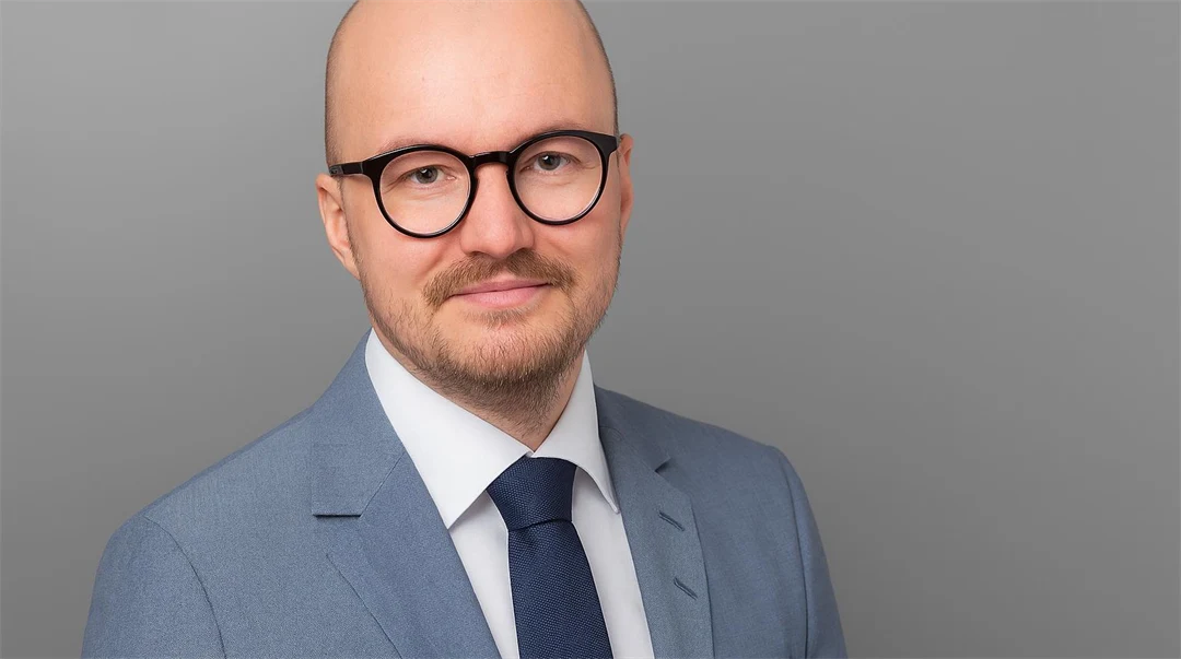 Man med glasögon och skägg, klädd i ljusgrå kavaj, vit skjorta och slips