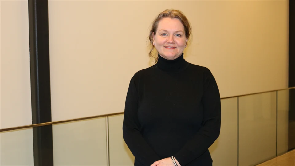 En kvinna med uppsatt brunt hår och svart polotröja i korridor med beiga väggar.