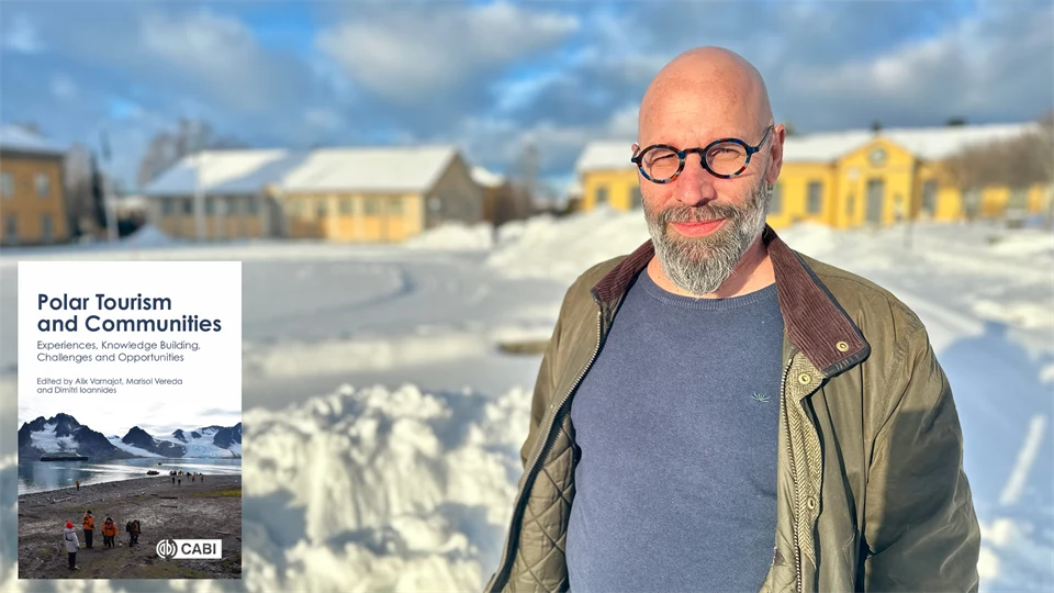 En man i glasögon, blå tröja och grön jacka står utomhus i ett snigt landskap med gula byggnader i bakgrunden. En bild på ett bokomslag är inklippt i bilden. 
