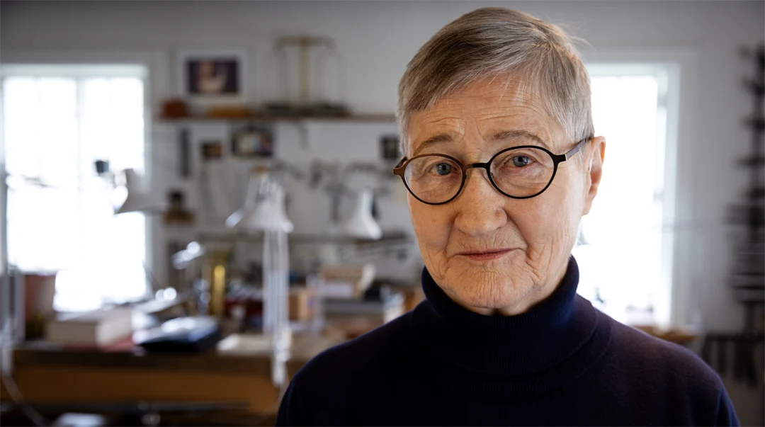 En kvinna med glasögon och kort hår tittar in i kameran. I bakgrunden ser man arbetsbänkar och hyllor.