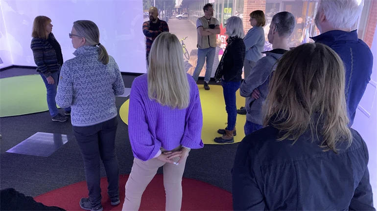En grupp människor står i ett rum med en stor skärm som visar en stadsmiljö.