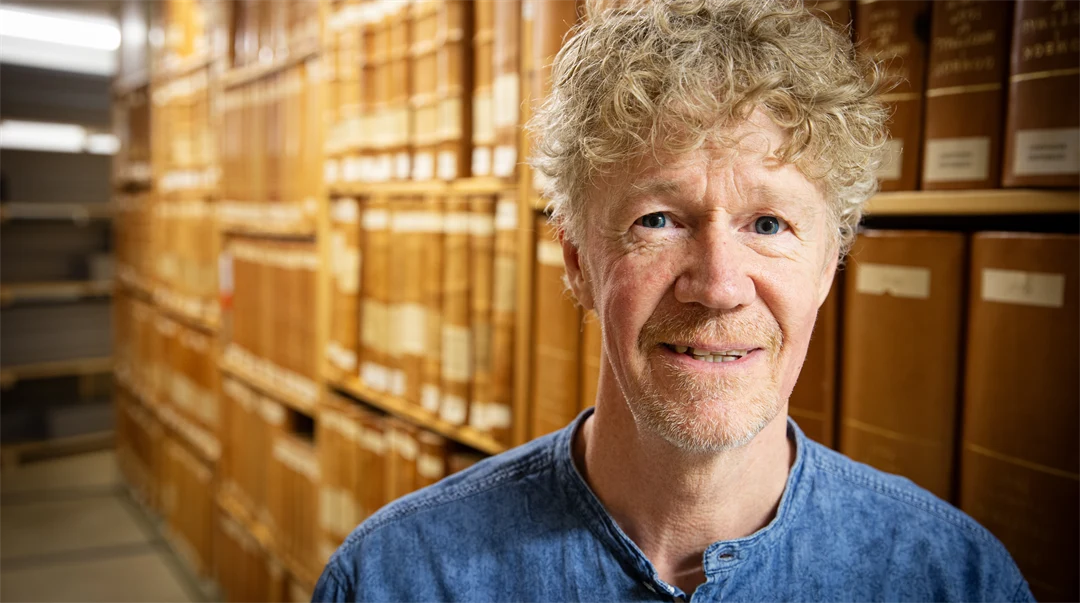 En man med blå skjorta tittar in i kameran. Bakom honom syns hyllor fyllda med gamla böcker. 