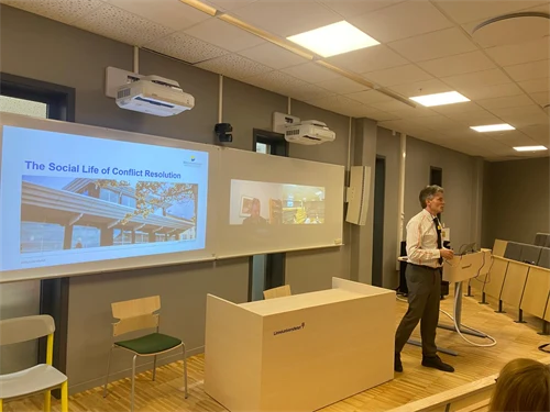 En man i vit skjorta, slips och mörka byxor står längst fram i en gradängsal med en skärm i bakgrunden.